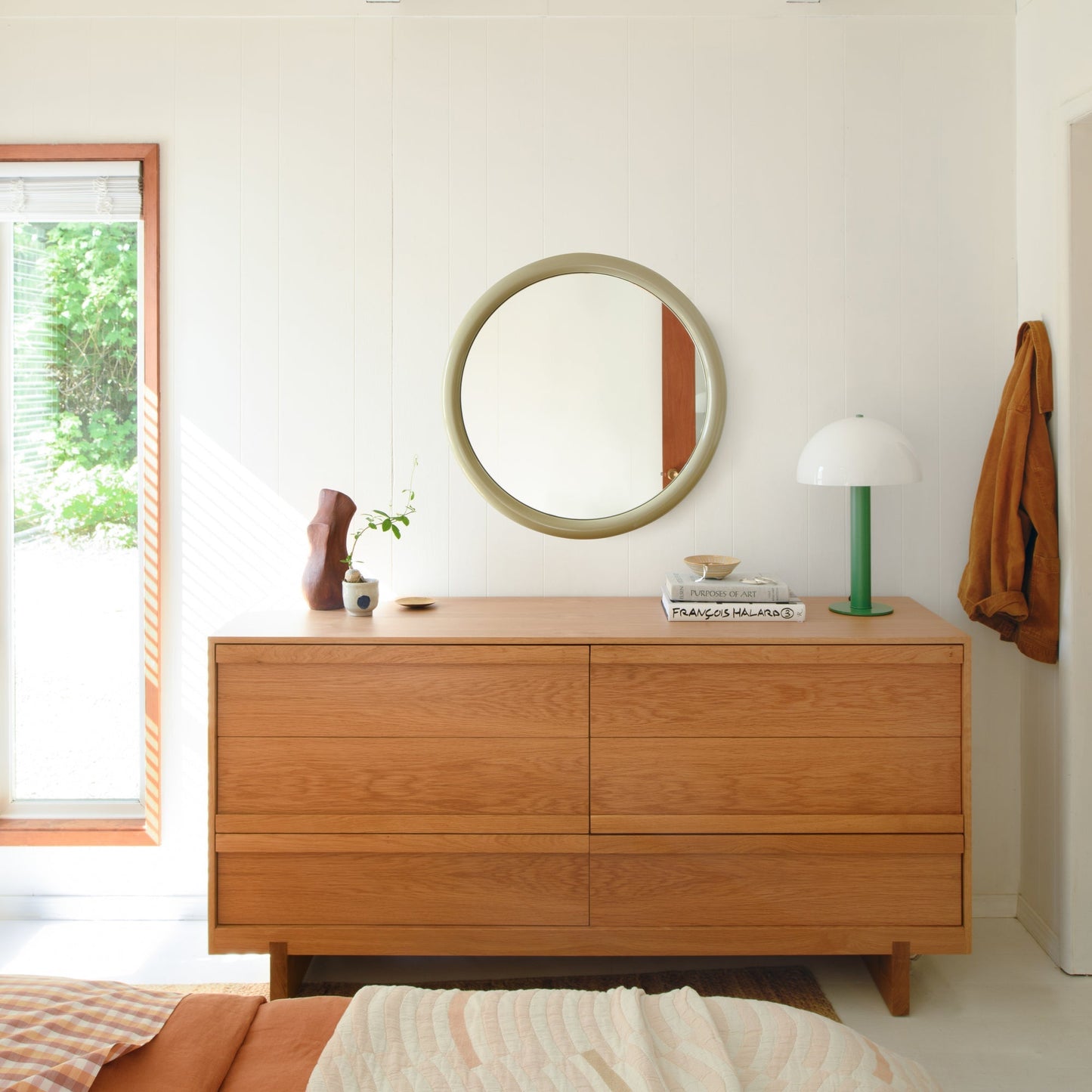 Front view of Forma mirror with olive-green round frame mounted above a wooden dresser in a bright, minimalist bedroom setting