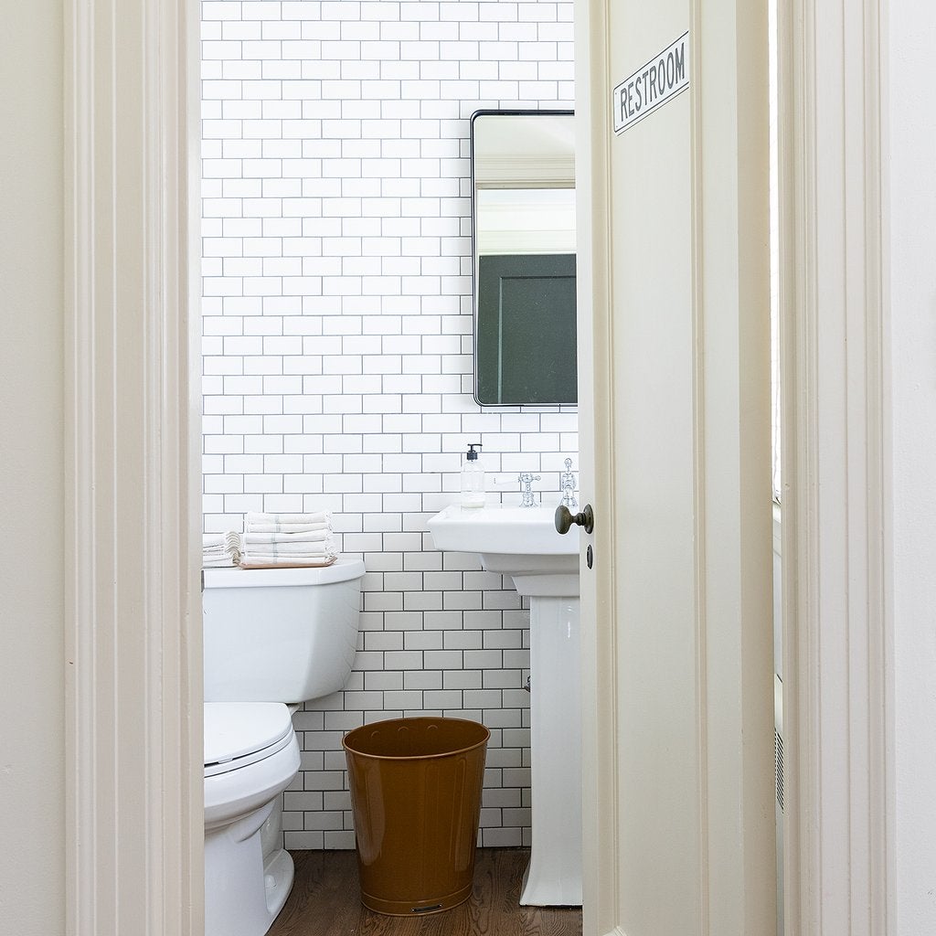 Rectangular wall mirror hung above a pedestal sink in a small white-tile bathroom, viewed through a half-open door