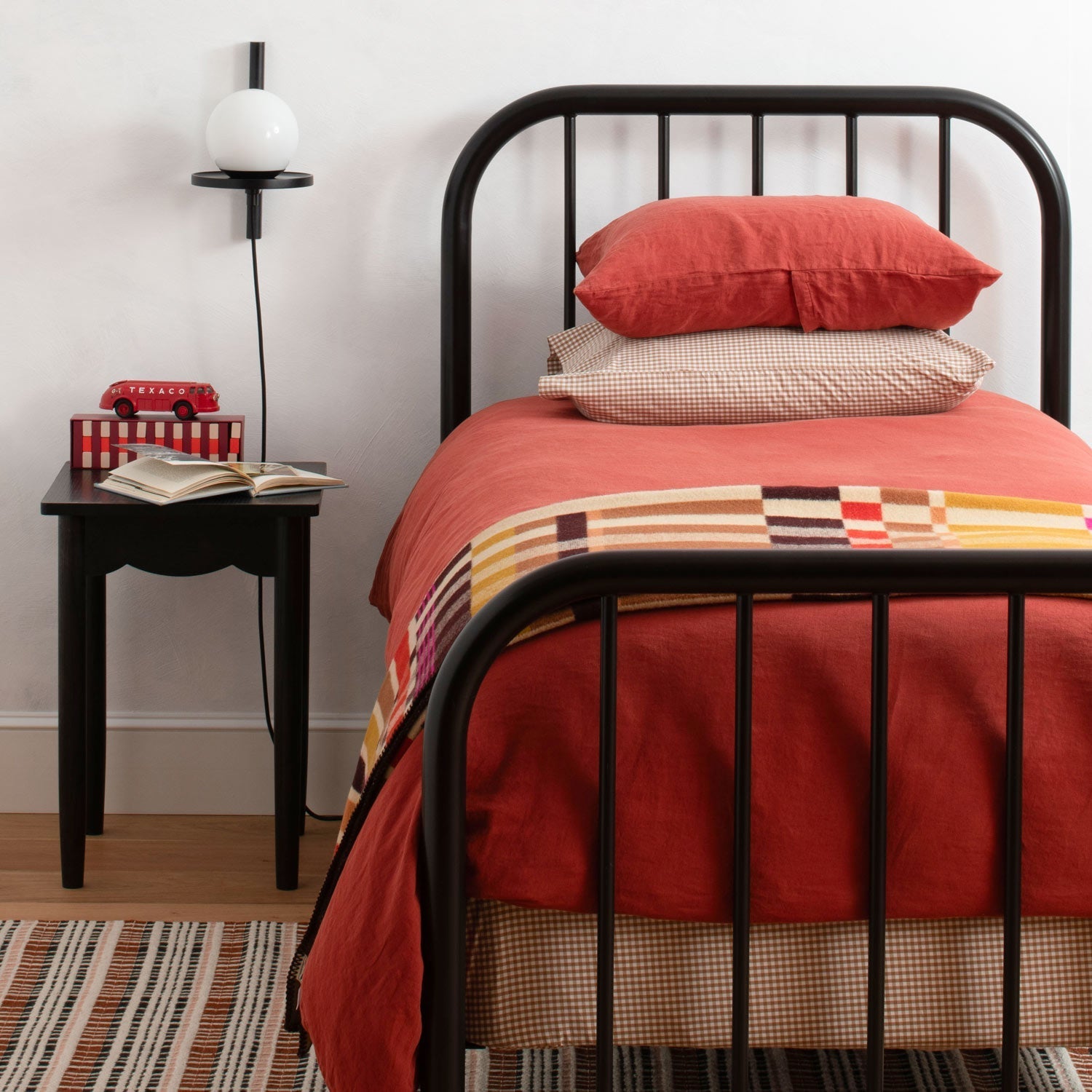 Stacked rust-colored linen pillow shams on a single bed with black metal frame, bedside table and wall sconce lamp