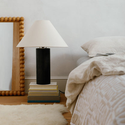 Bobbin mirror leaning against a wall beside a bed with a black cylindrical lamp on stacked books and neutral linen bedding