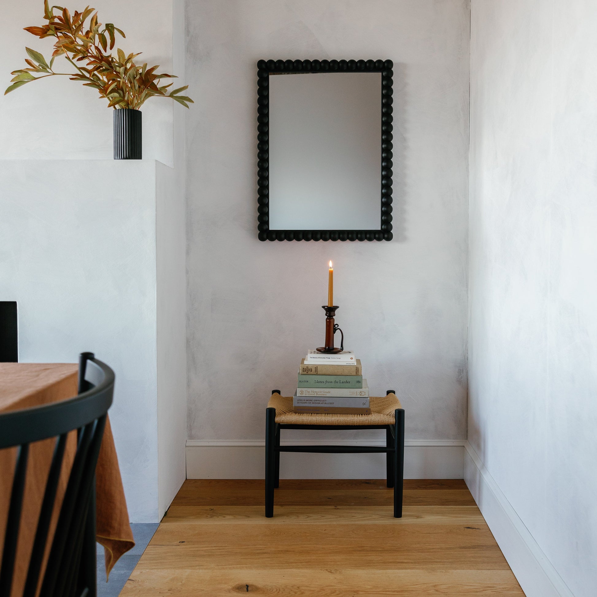 Front view of Bobbin Mirror showing black beaded frame against a white plaster wall with a lit candle and vase