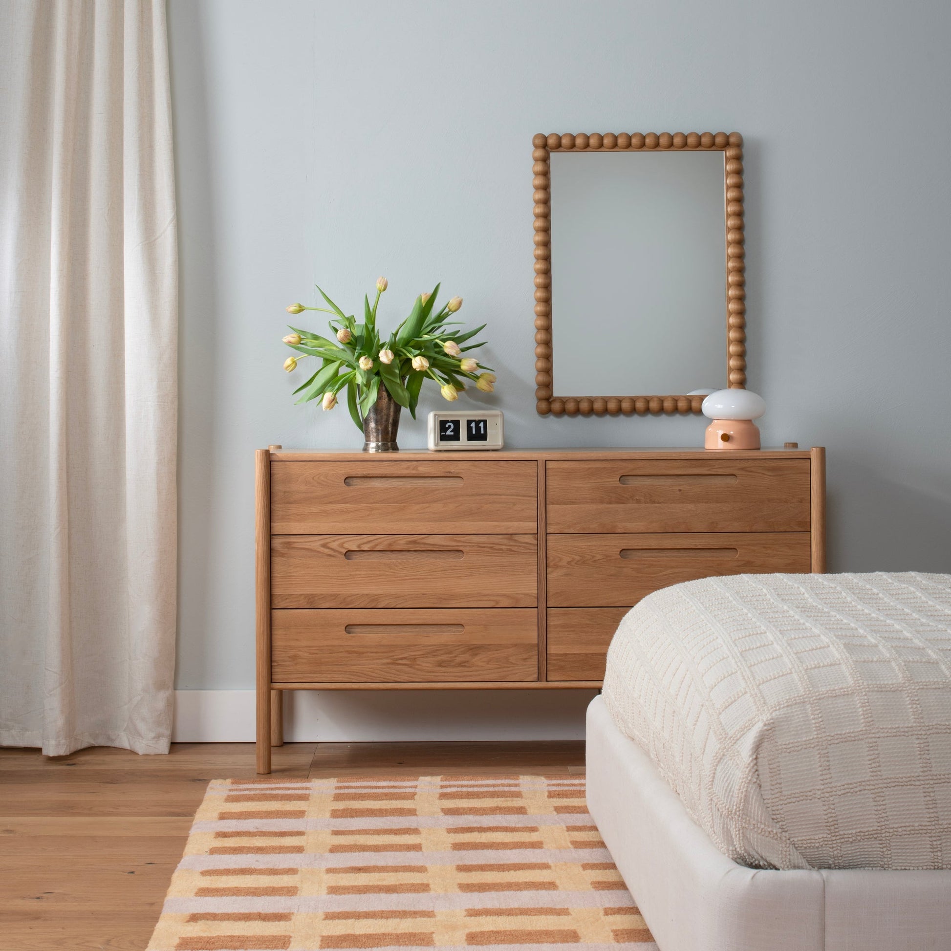 Straight-on view of Bobbin Mirror showing its wooden beaded frame hung above a wooden dresser with tulips, a flip clock, and a lamp