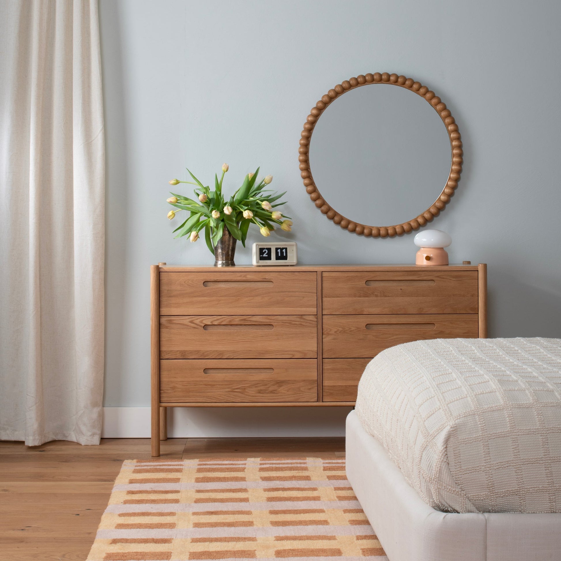 Hanging 30-inch Bobbin Mirror in Ash showing beaded wooden frame above a wood dresser styled with tulips and a lamp