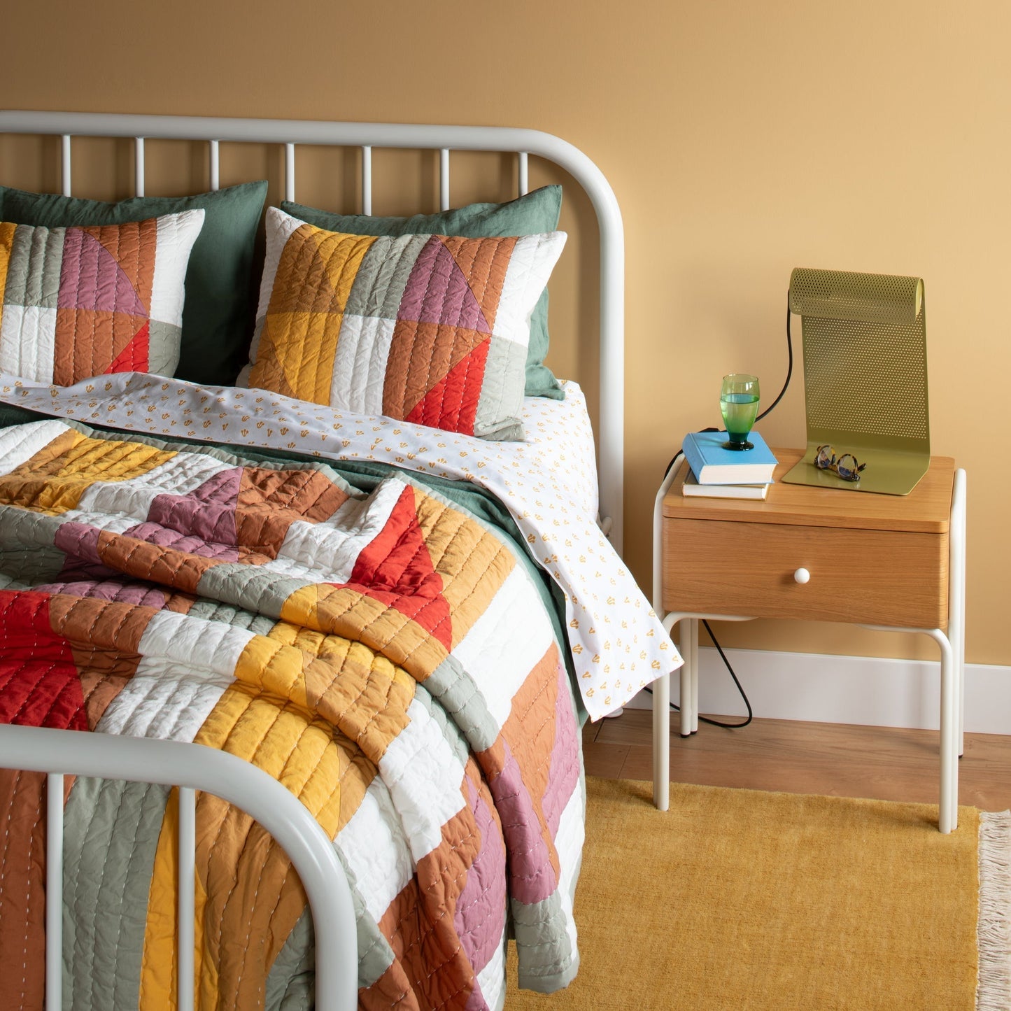 Mesa Wool Rug peeking from under a white metal bed in a warm bedroom with patchwork quilt and wooden nightstand