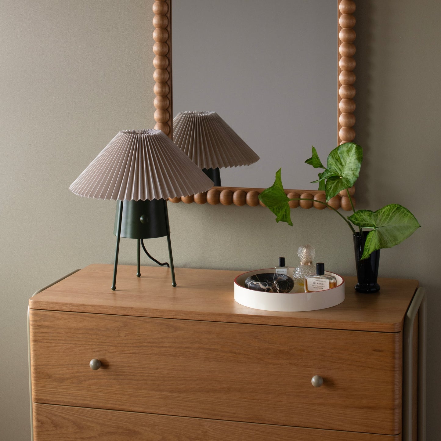 Partial front view of Bobbin Mirror showing beaded wood frame, reflected pleated lamp, and a potted plant on a dresser