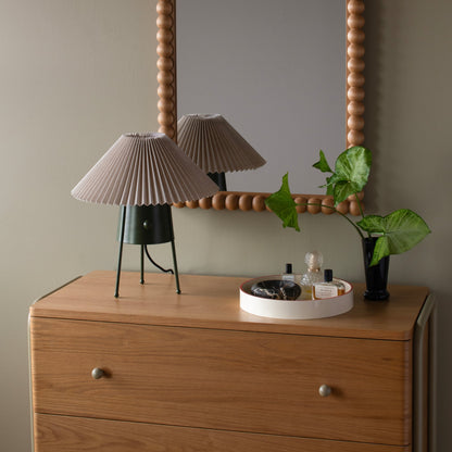 Partial front view of Bobbin Mirror showing beaded wood frame, reflected pleated lamp, and a potted plant on a dresser