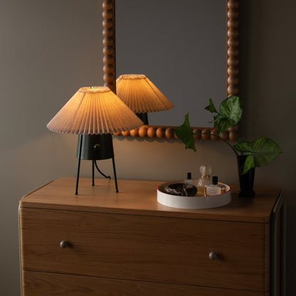 Bobbin mirror reflecting a pleated table lamp and trailing plant above a wooden dresser in warm ambient light