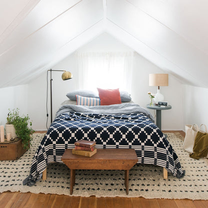 Bed styled with Winter + Summer cotton coverlet (navy geometric) layered with pillows in a bright airy attic bedroom