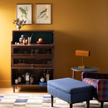 Campus Wool Rug shown under a blue ottoman and plaid chair in a warm living room with a mustard wall and wooden cabinet