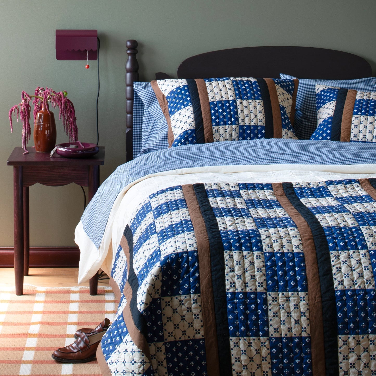 Plaid wool reversible rug shown under a bed in a bedroom with a blue patchwork quilt and wooden nightstand