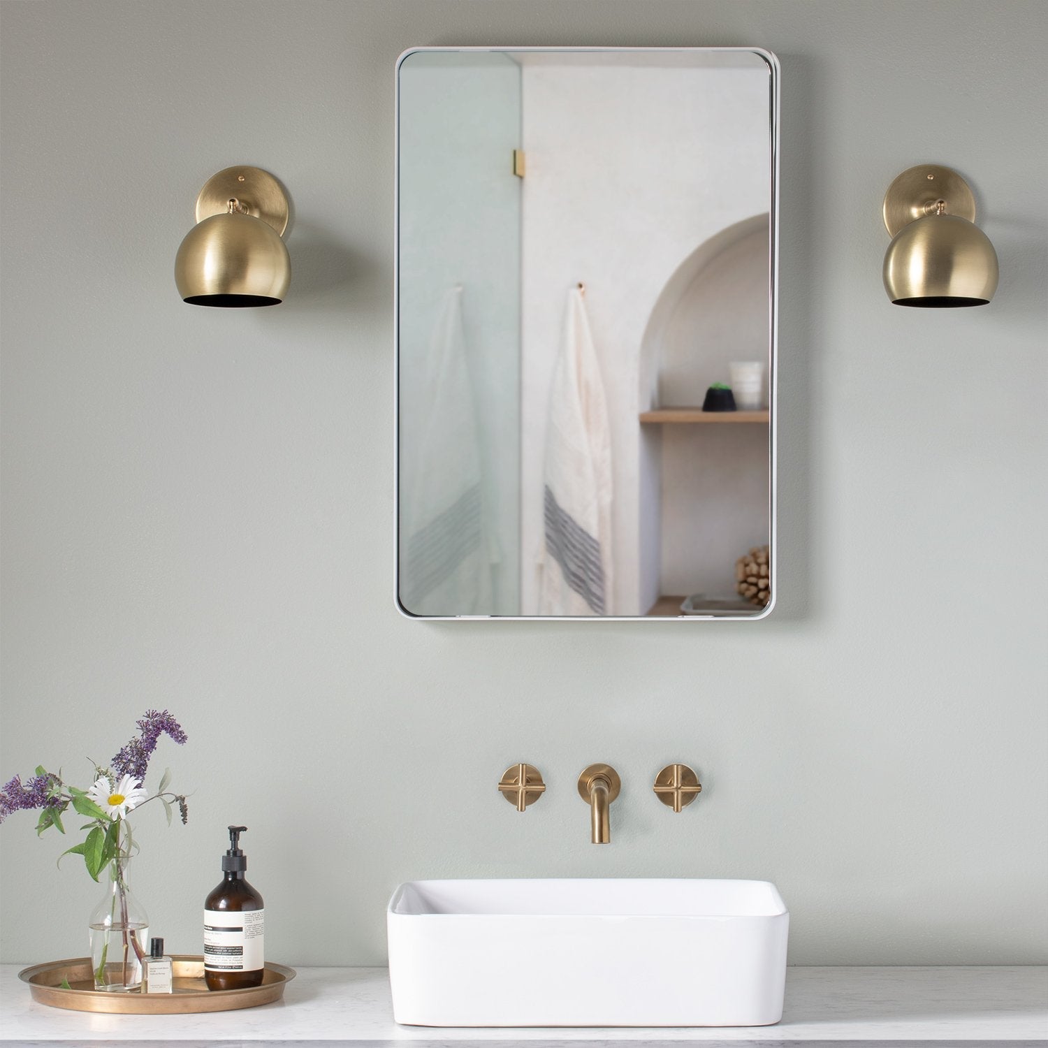 Front view of Leo mirror showcasing thin rounded metal frame with polished edge, mounted above a sink with brass sconces