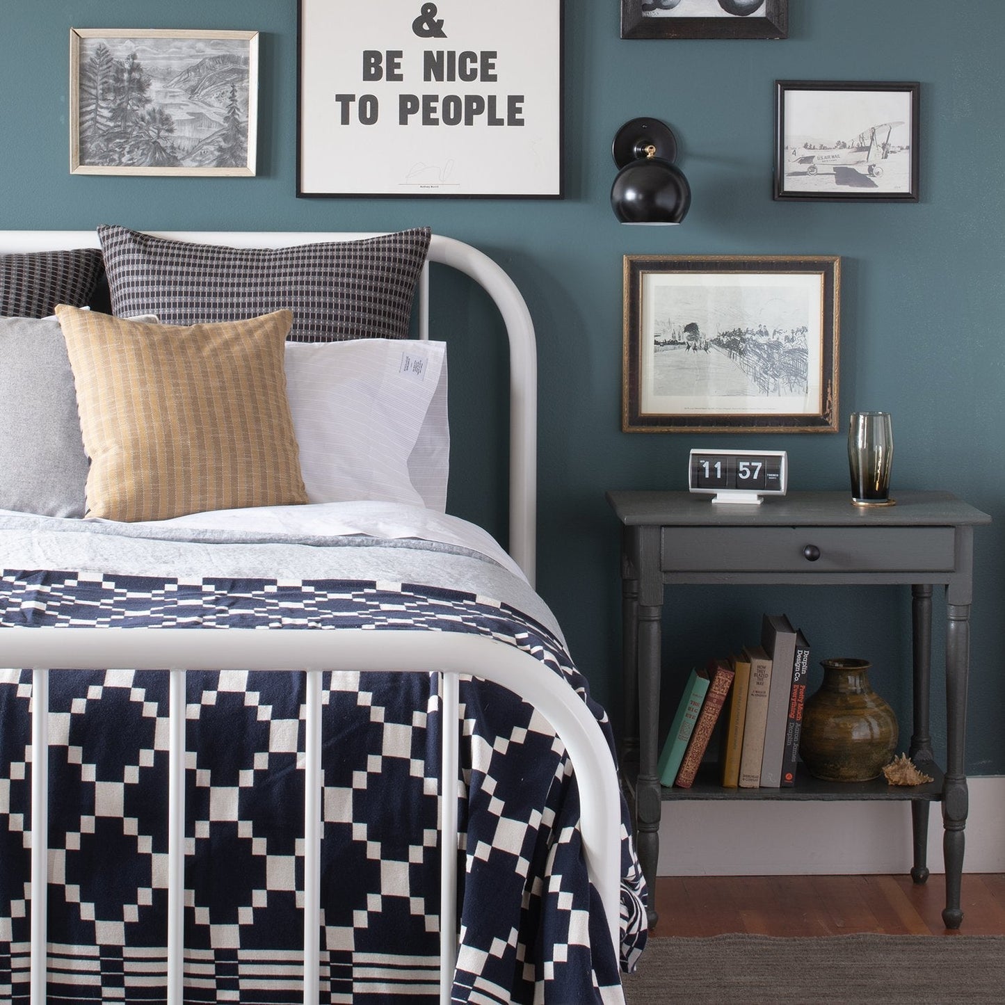 Winter + Summer Cotton Coverlet in navy with white draped on a white metal bed, geometric pattern, gray nightstand, framed print reading ‘Be nice to people’