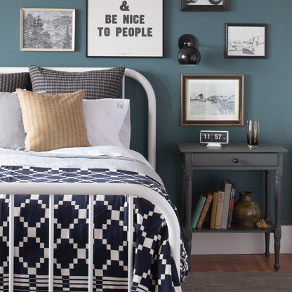 Winter + Summer Cotton Coverlet in navy with white draped on a white metal bed, geometric pattern, gray nightstand, framed print reading ‘Be nice to people’
