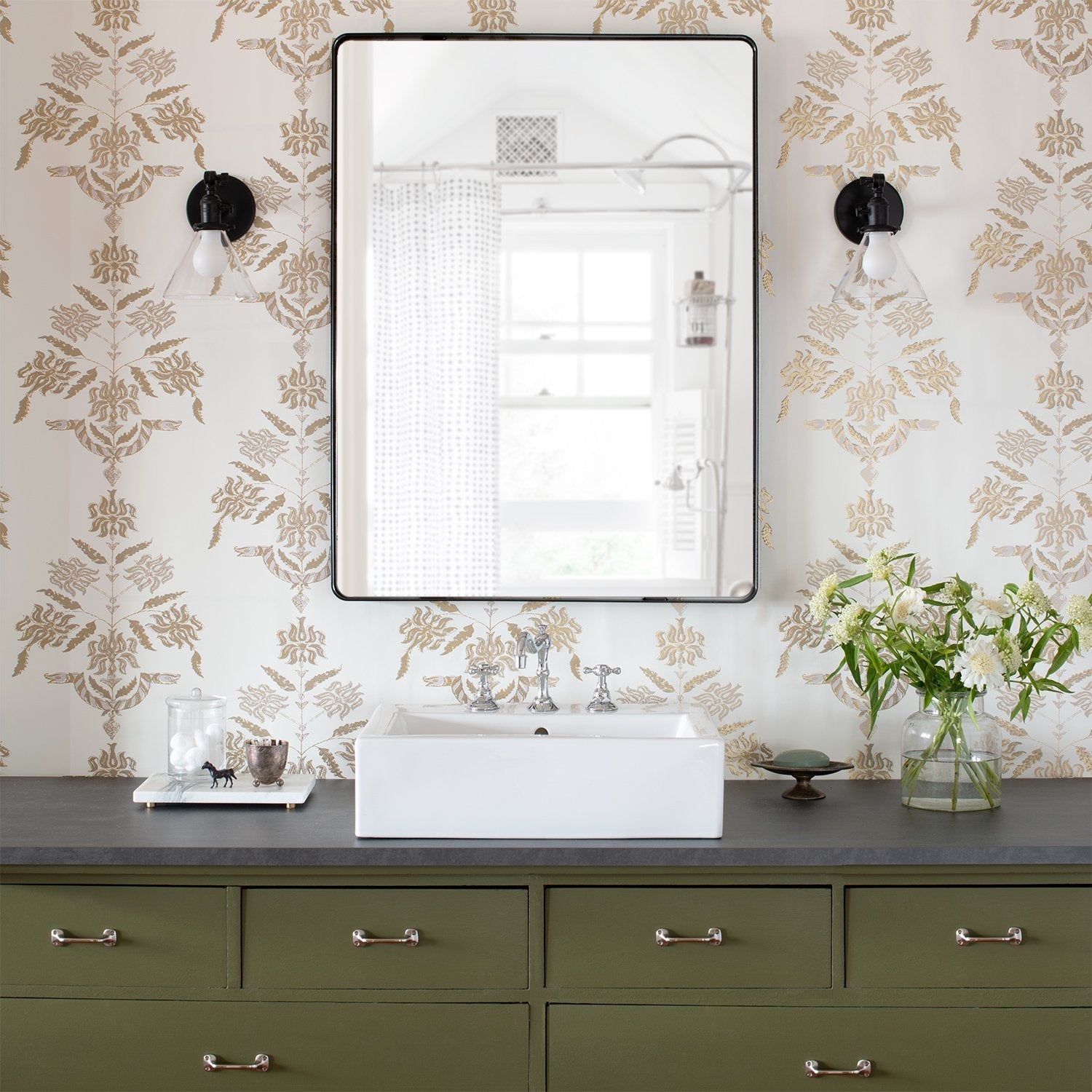 Rectangular Leo mirror above a white vessel sink in a bathroom with gold damask wallpaper, black glass sconces, and an olive green vanity
