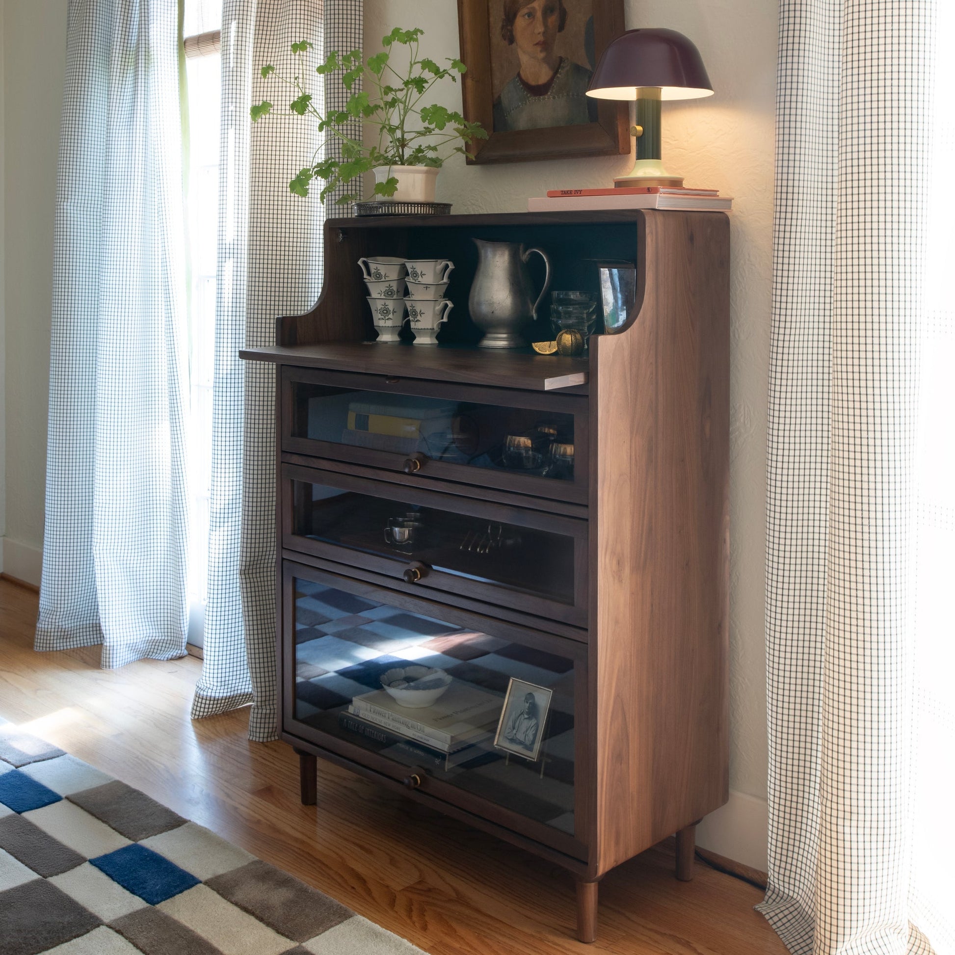 Sunlit corner with Campus Wool Rug on a hardwood floor beside a wooden side cabinet, potted plant, and table lamp