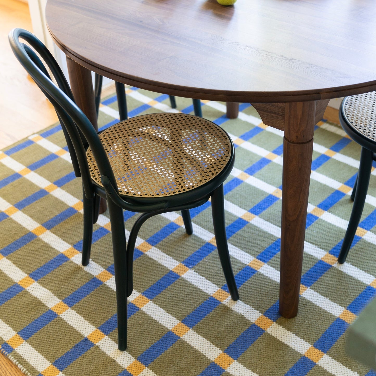 Plaid wool reversible rug beneath a wooden dining table with black cane seat chairs on olive blue white and yellow plaid