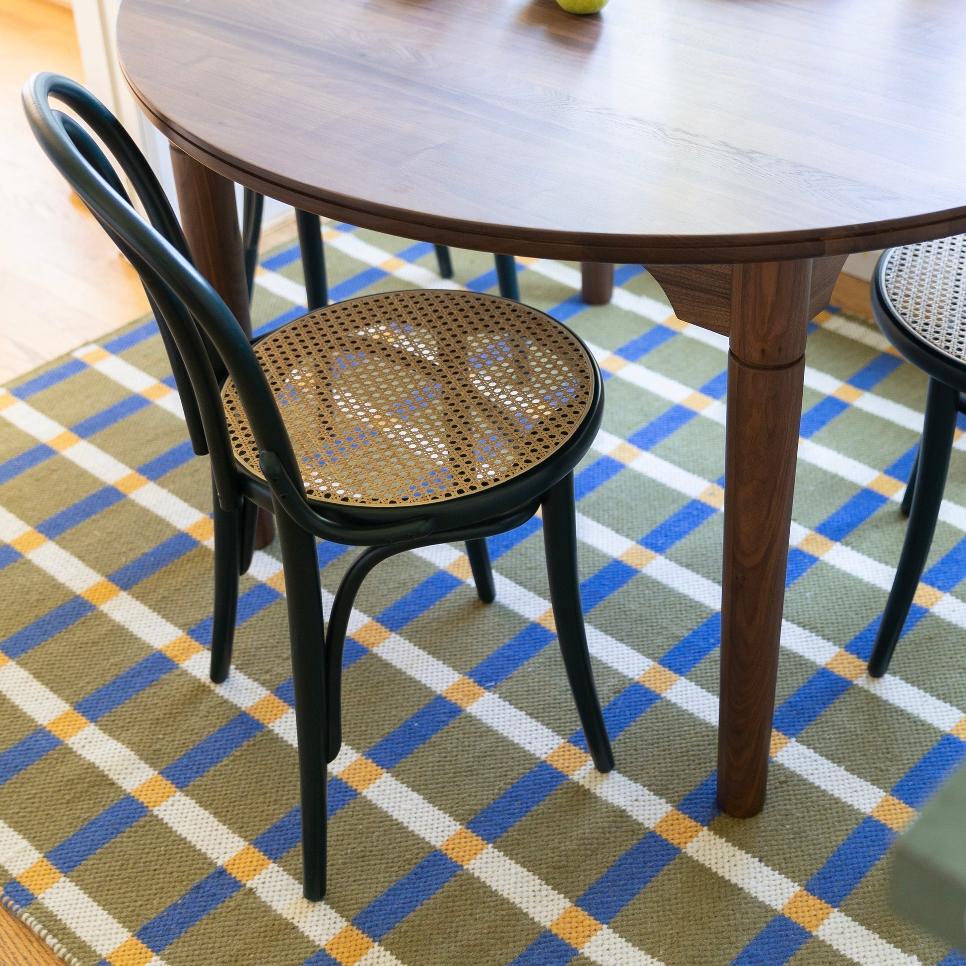 Plaid wool reversible rug beneath a wooden dining table with black cane seat chairs on olive blue white and yellow plaid