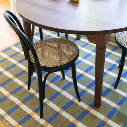 Plaid wool reversible rug beneath a wooden dining table with black cane seat chairs on olive blue white and yellow plaid