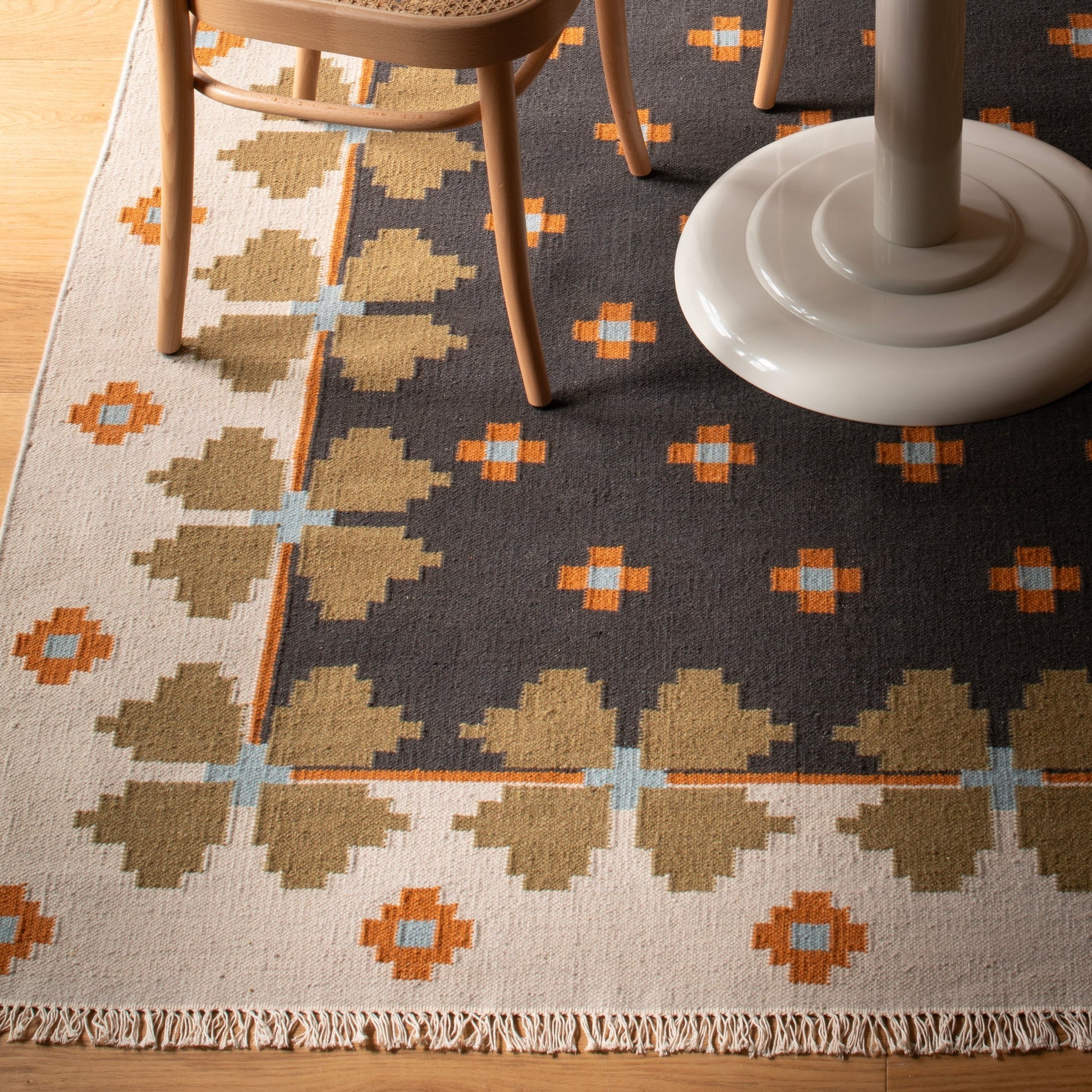 Close-up of Flora Wool Flatweave Rug showcasing its wool flatweave folk‑floral pattern, fringed edge, and two-tone border under chair legs on a wood floor