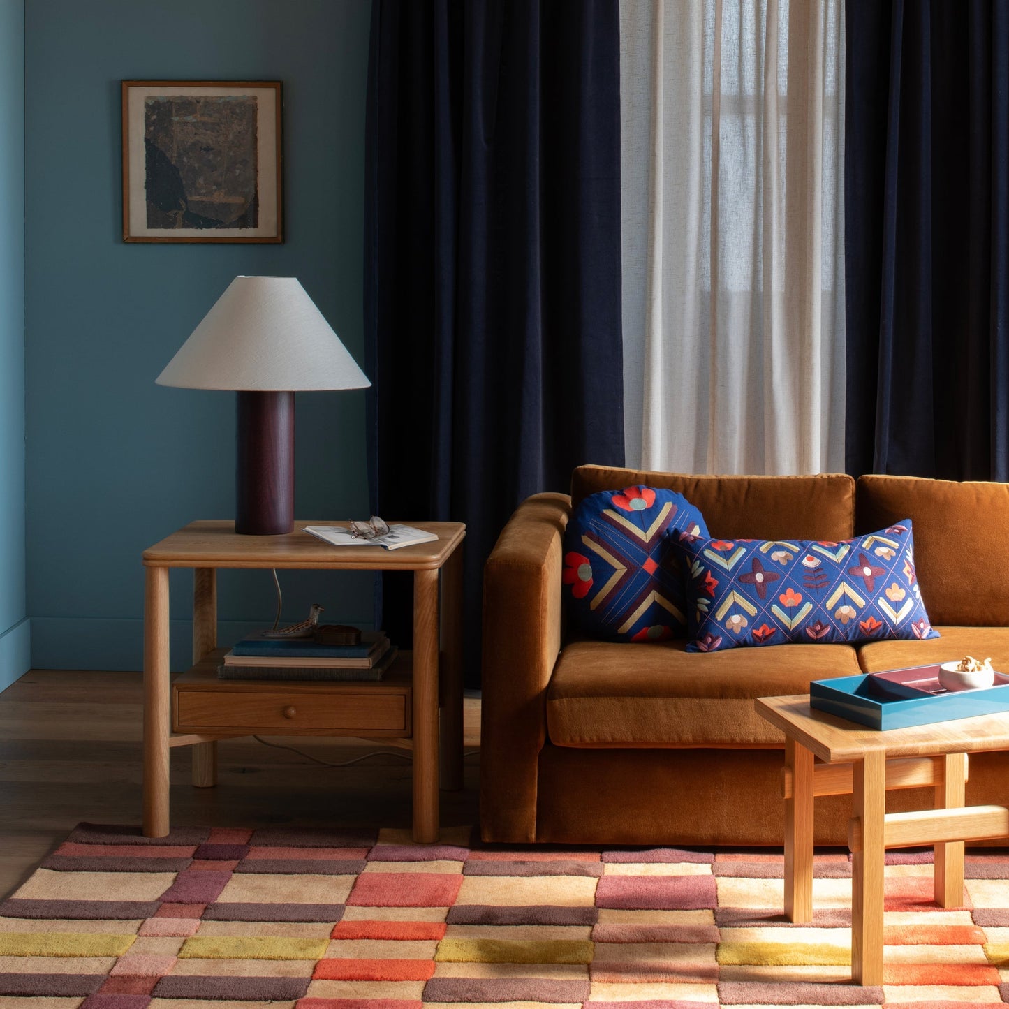 Campus Wool Rug shown on living room floor with plaid-inspired graduated squares in reds, pinks, and yellow beside a brown velvet sofa and wooden side table