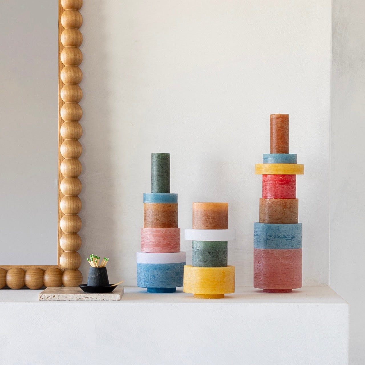 Beaded wood bobbin-framed mirror hangs above a white mantel with three stacked multicolor pillar candles and a black dish