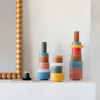 Beaded wood bobbin-framed mirror hangs above a white mantel with three stacked multicolor pillar candles and a black dish