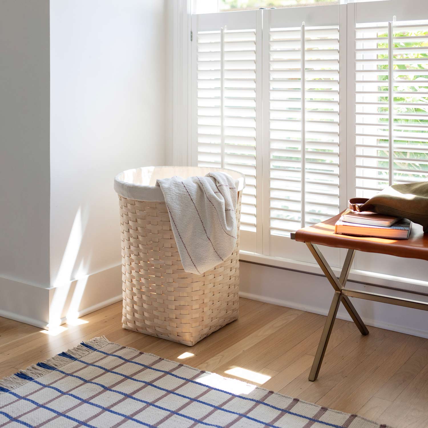 Cotton grid flatweave rug laid on hardwood floor by a wicker laundry basket and leather bench in a sunlit room