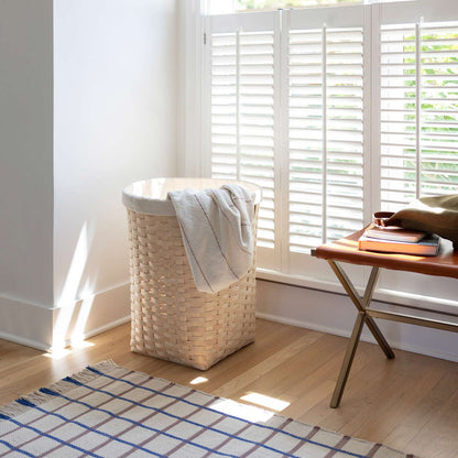Cotton grid flatweave rug laid on hardwood floor by a wicker laundry basket and leather bench in a sunlit room