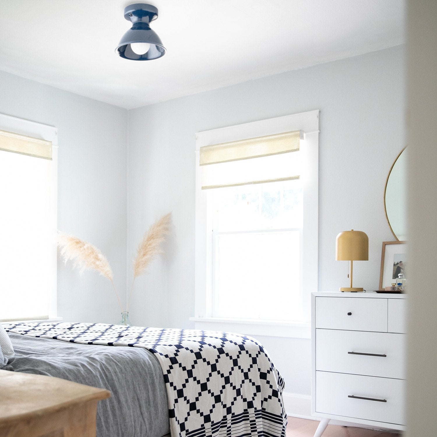 Sunlit bedroom showing a navy-and-white geometric cotton coverlet layered over gray bedding beside a white dresser with a gold lamp