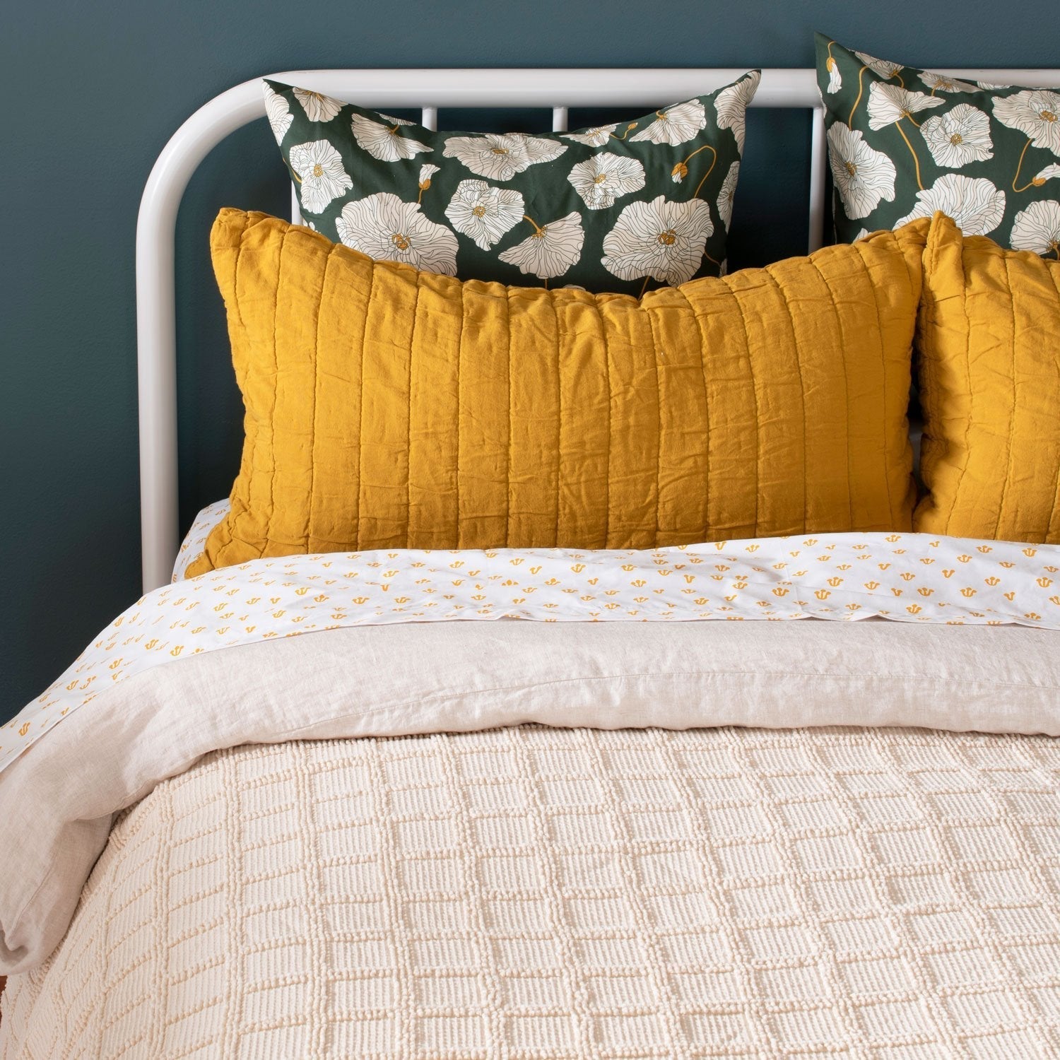 Blooming Field shams displayed on a bed with mustard quilted pillows, cream bedding, and a white metal headboard