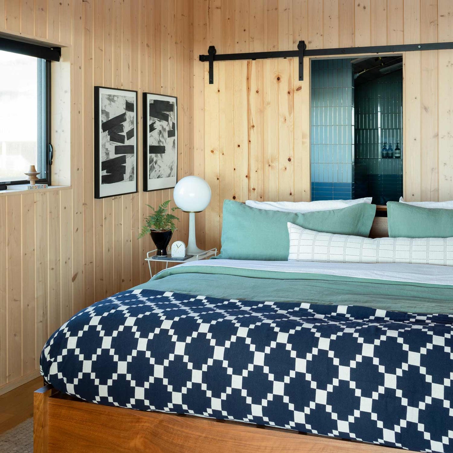 Bed dressed with Winter + Summer cotton coverlet in navy and white, layered with seafoam green pillows in a wood-paneled bedroom