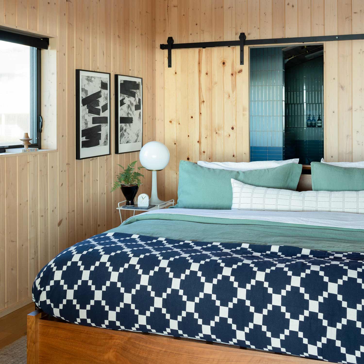Bed dressed with Winter + Summer cotton coverlet in navy and white, layered with seafoam green pillows in a wood-paneled bedroom