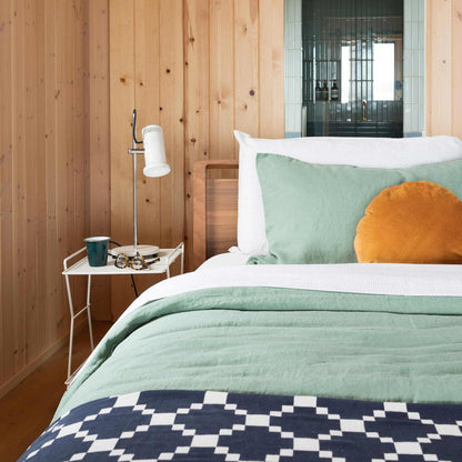 Bed dressed with a light green cotton coverlet layered with white sheets and a navy geometric throw in a wood-paneled bedroom