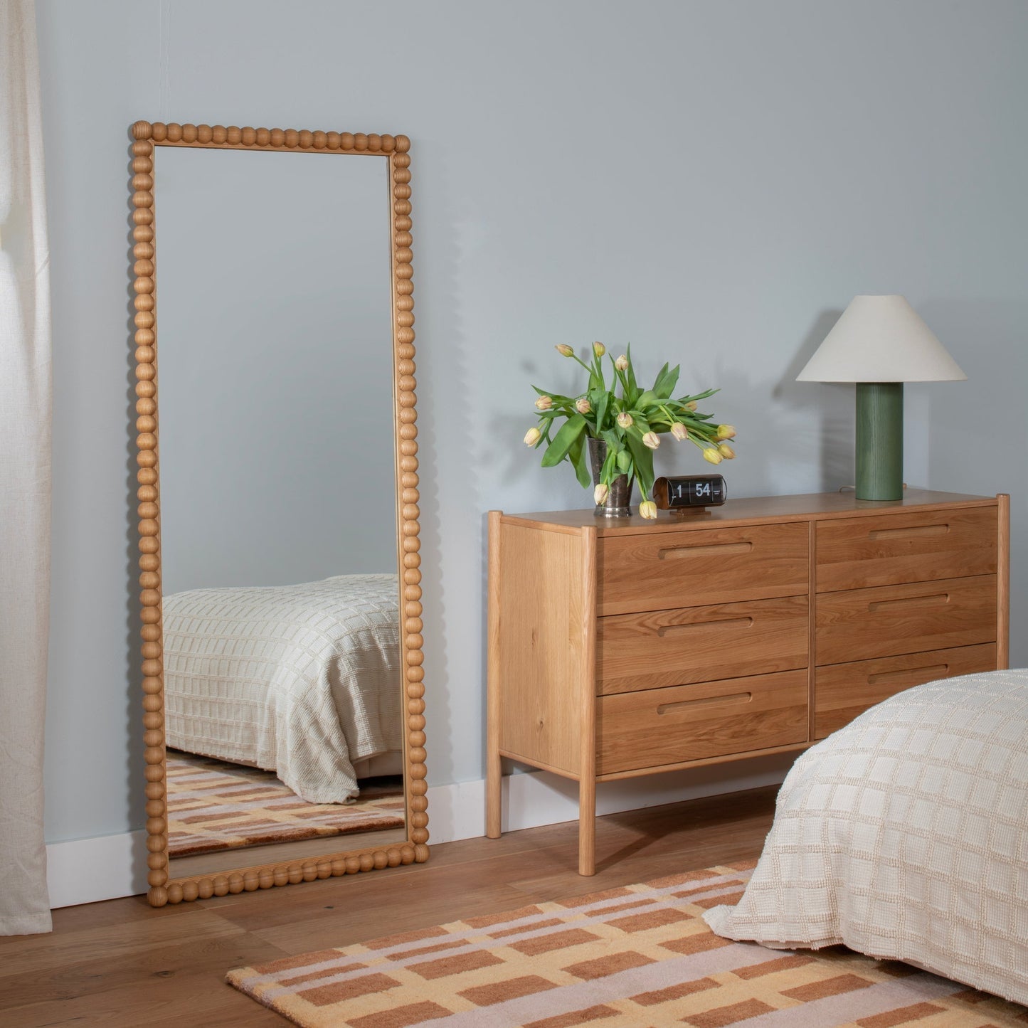 Full-length view of Bobbin Mirror showcasing beaded ash wood frame and mirrored glass against a pale blue wall above an oak dresser