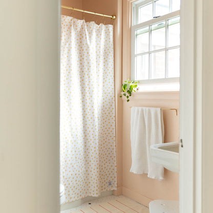Shower curtain with small honey yellow thistle print hanging from brass rod in sunlit peach bathroom with towel on gold bar
