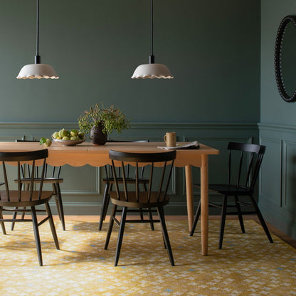 Bobbin mirror on a green paneled dining room wall above a scalloped edge wooden table with black chairs and pendant lights