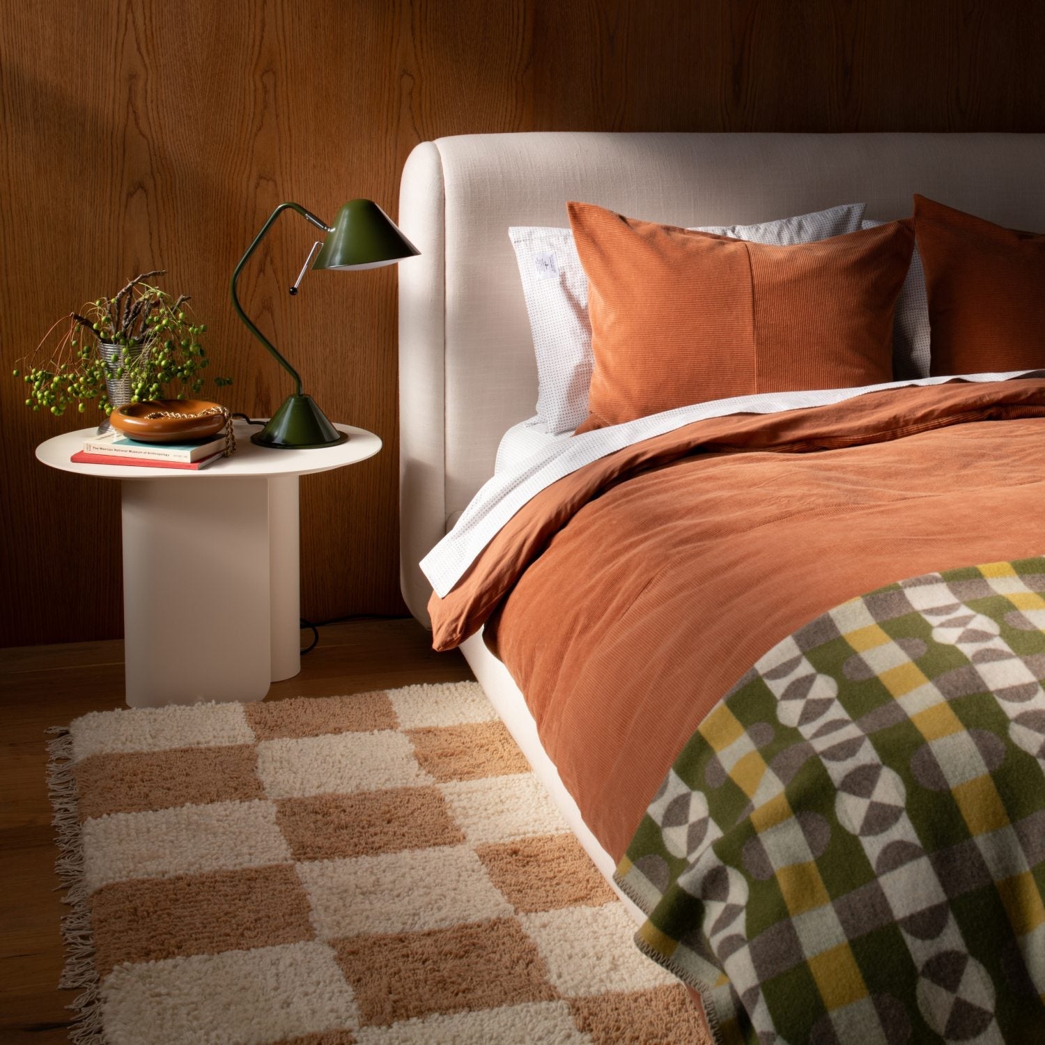Bed with rust orange linen duvet and pillows beside a white round nightstand with a green task lamp on a checkered rug