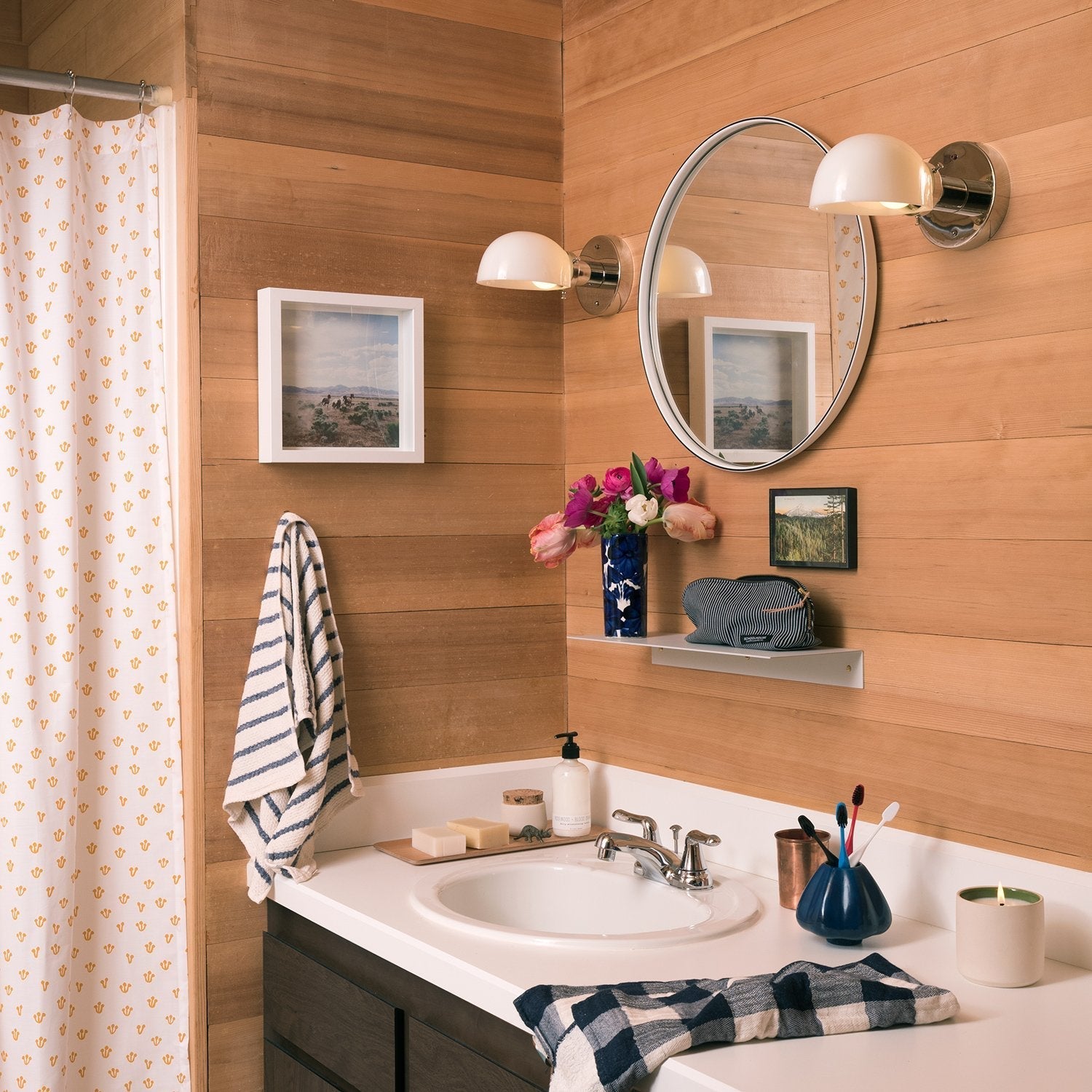 White shower curtain with small mustard-yellow thistle print hanging in a wood-paneled bathroom beside a sink and round mirror