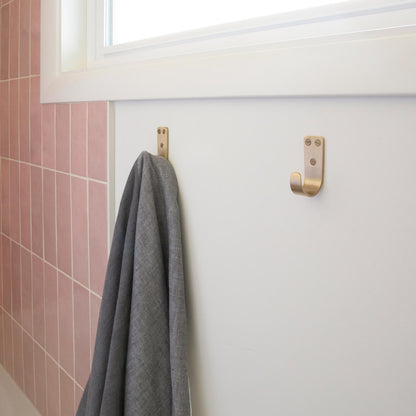 Close-up of Thomas Hook in natural brass showing three mounting holes and curved hook on white wall with pink tile and gray towel