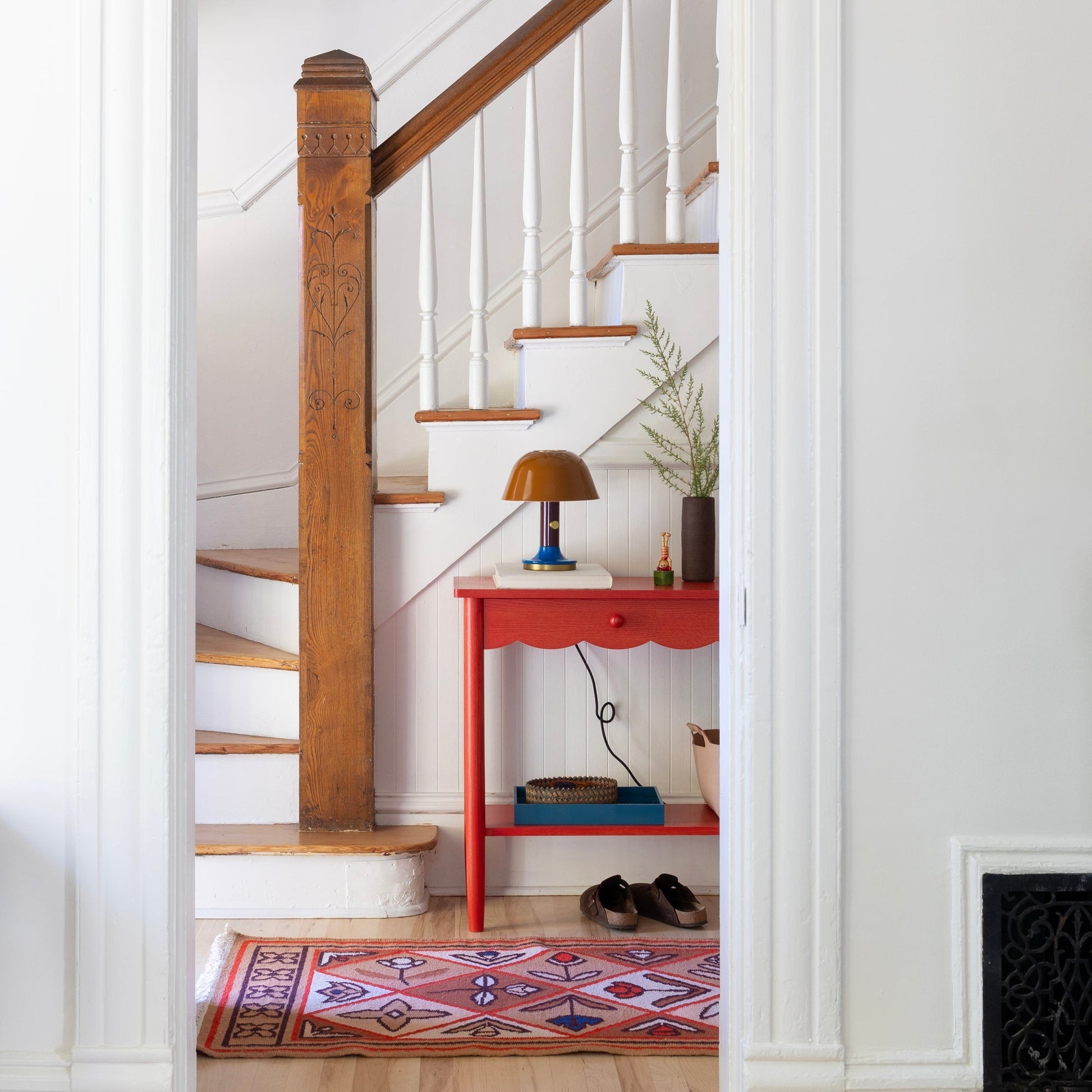 Linnea wool flatweave rug in an entryway beside a carved wood newel post, white staircase, and red side table with lamp