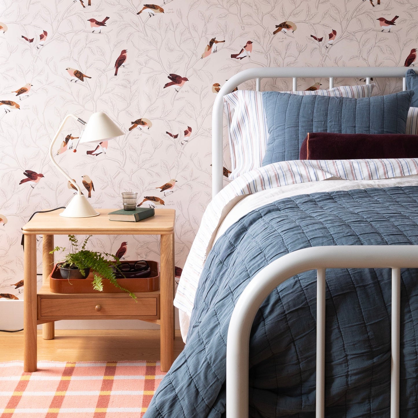 Plaid wool rug on a hardwood floor in a cozy styled bedroom with a white metal bedframe, striped bedding, bird-print wallpaper, and a wooden nightstand
