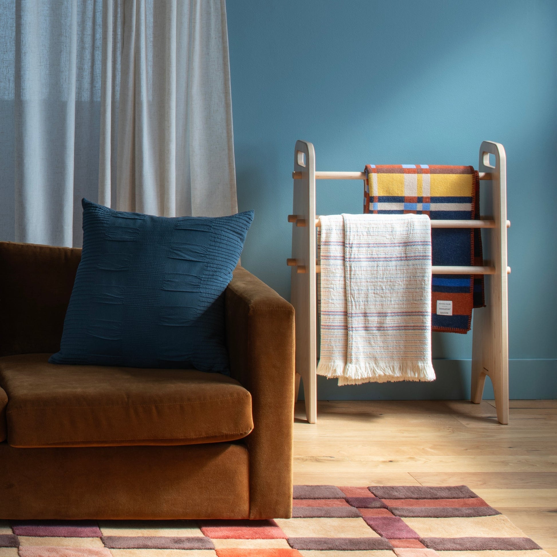 Living room scene with brown sofa, blue pillow, wooden blanket ladder, and Campus Wool Rug visible on hardwood floor against a blue wall