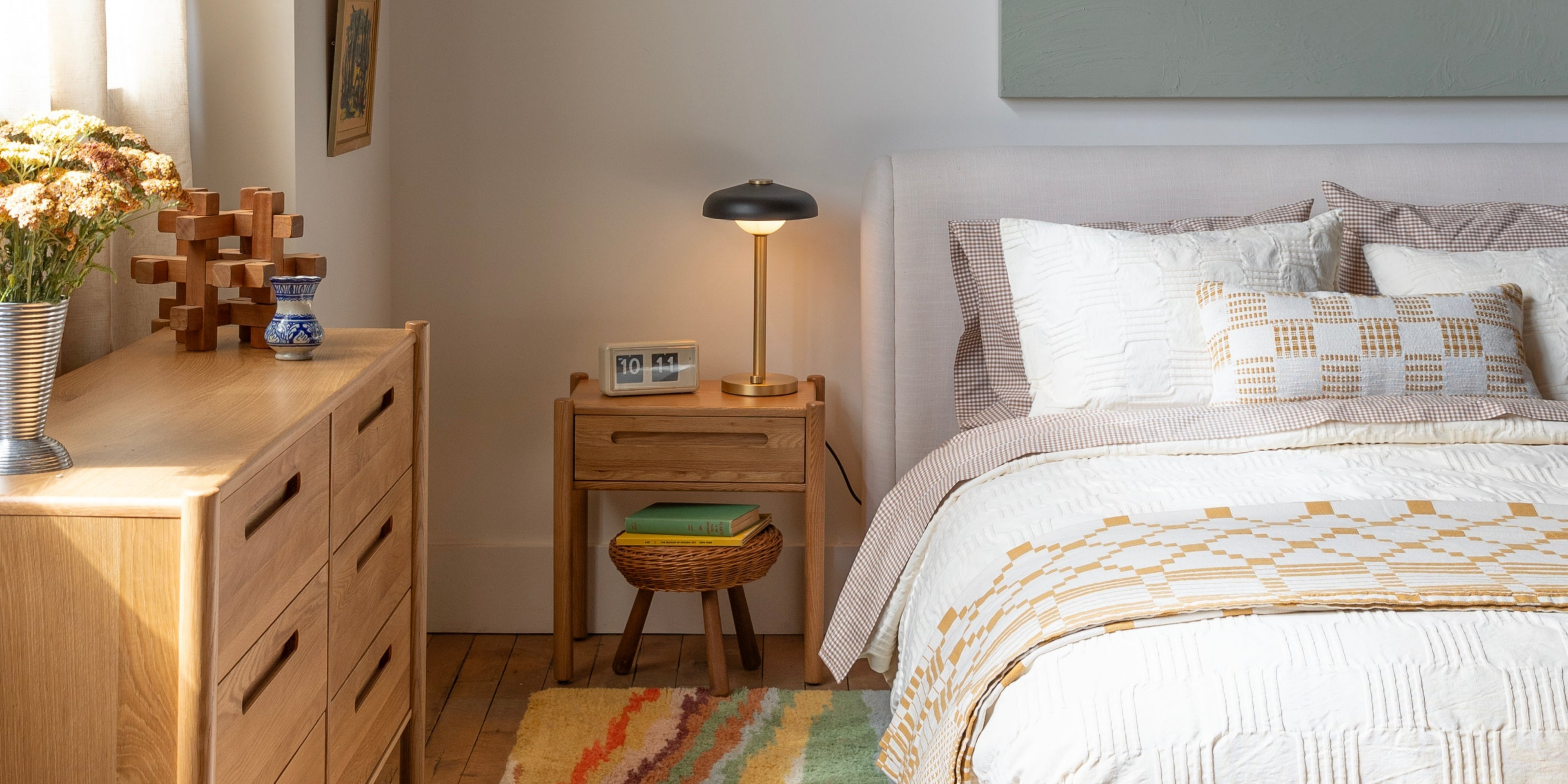 Sunlit bedroom with an upholstered bed dressed in a white quilt and patterned pillows, wooden nightstand with a brass lamp and dresser