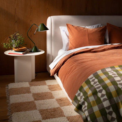 Yves cast metal catch-all on a white bedside table beside a green lamp in a bedroom vignette with rust-colored bedding and wood-paneled wall