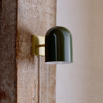 Side view of Allegheny 6-inch plug-in sconce showing glossy green cylindrical shade, brass wall plate, and fabric-wrapped cord on weathered wood