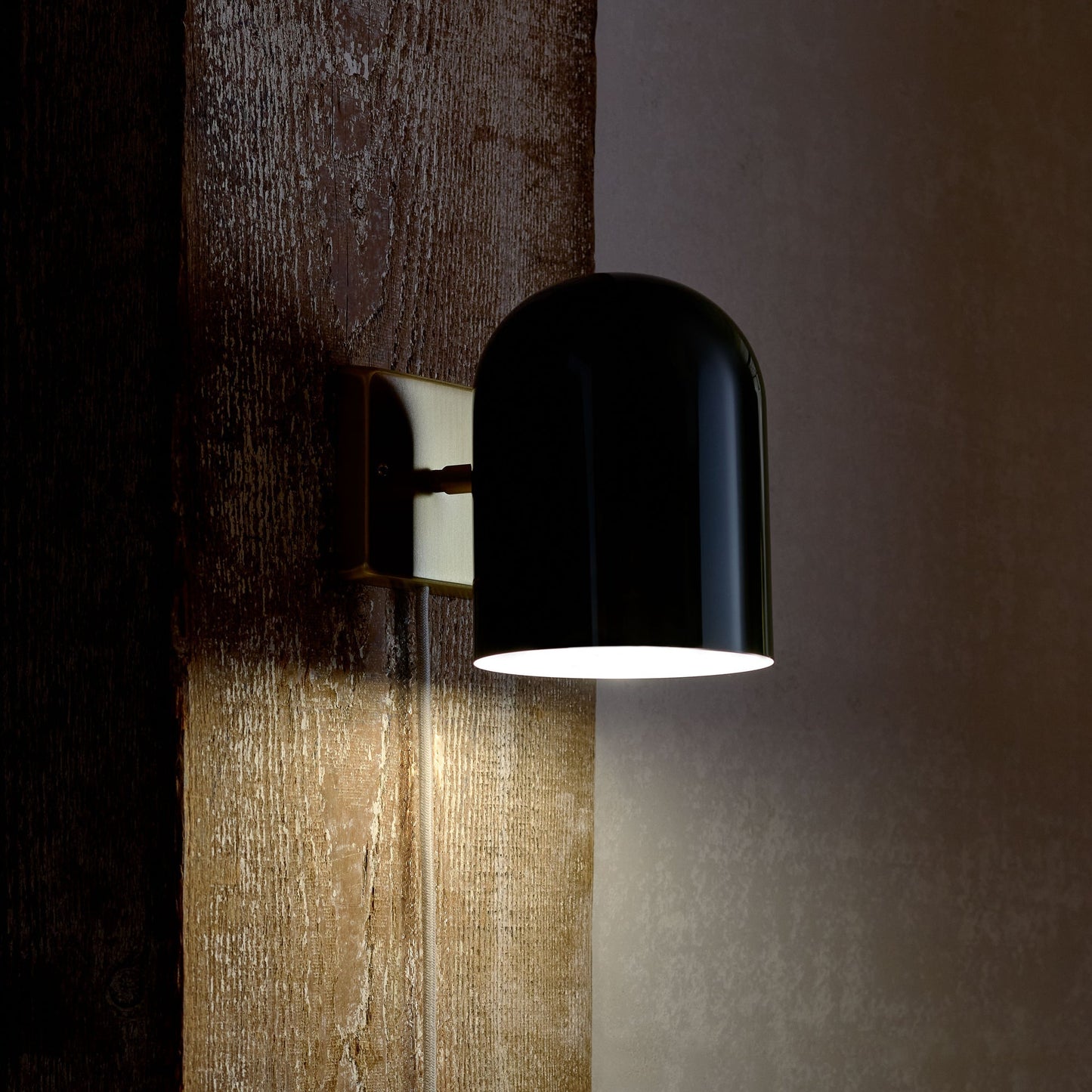 Side view of Allegheny 6” plug-in sconce showing glossy black cylindrical shade and brass wall plate on a textured wood wall