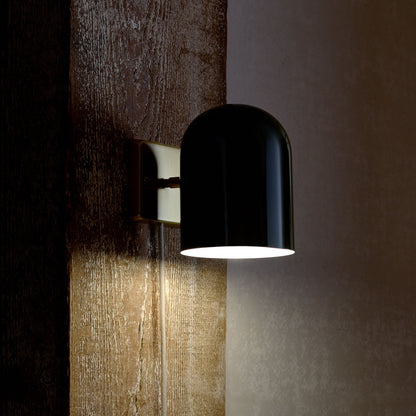Side view of Allegheny 6” plug-in sconce showing glossy black cylindrical shade and brass wall plate on a textured wood wall