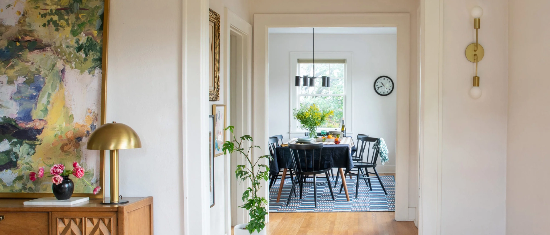 Dining room arranged for a meal seen through a hallway, black chairs around a table with yellow flowers and a brass lamp on a sideboard