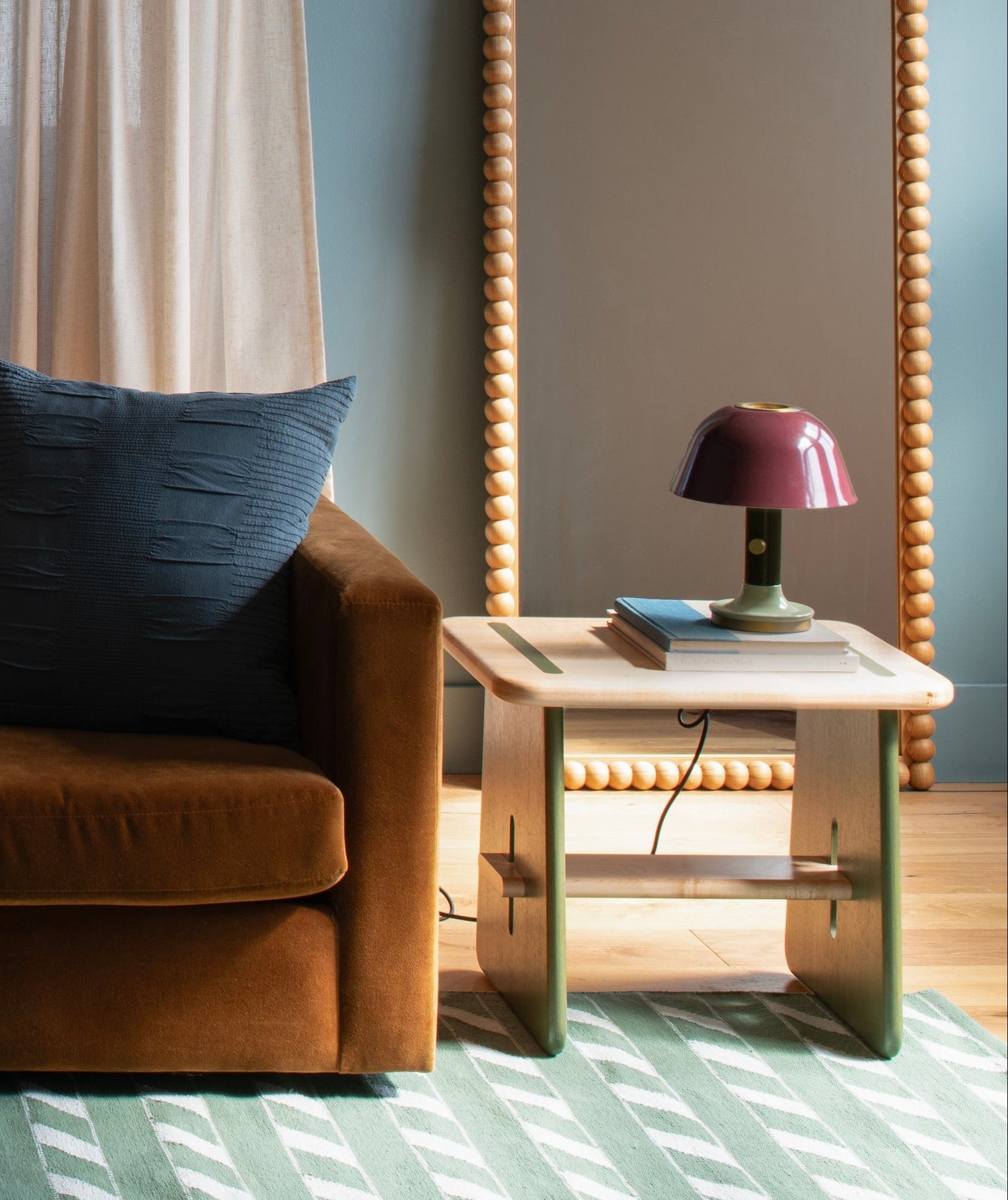 Rust-colored sofa beside a light wood side table with green accents, stacked books and a plum table lamp in a living room with a beaded mirror and patterned rug