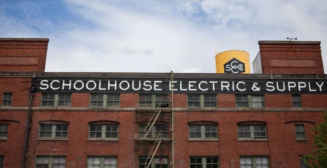 Schoolhouse factory building exterior showing painted sign reading SCHOOLHOUSE ELECTRIC & SUPPLY and yellow water tower under cloudy sky